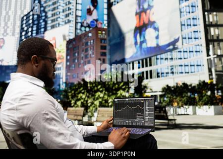 Black businessman sitting on a bench monitoring stock market trends on laptop, working remotely on laptop from a bench outdoor. Sitting in small park downtown and analyzes funds. Stock Photo