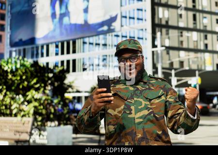 Professional smiling military man reading satisfying good news on mobile phone, cheering in the city center and feeling happy with his accomplishment. Honor, loyalty and security. Stock Photo