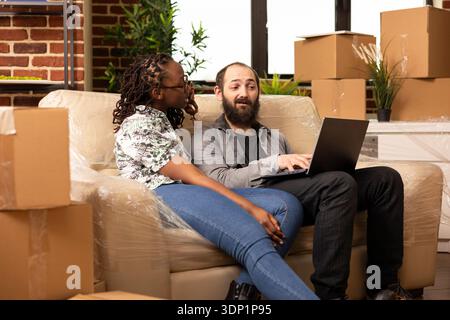 Multiethnic spouses relax on couch in new apartment, browsing laptop for furniture ideas. African american woman and caucasian man take break from unpacking, watching videos together on device. Stock Photo