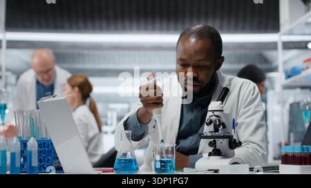 Materials scientist pipetting blue solution into test tube to analyze it. Microscopist transferring chemical agent to glass flask prepare for immediate spectroscopic analysis, camera B Stock Photo
