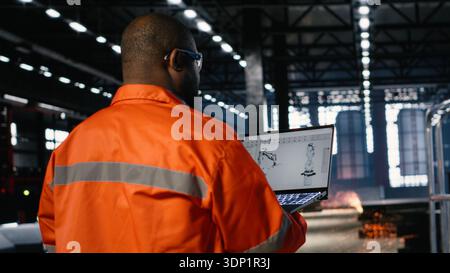 Worker using automation software connected to robot machinery via laptop, surrounded by metal equipment and fabrication tools. Industrial environment highlights engineering. Stock Photo