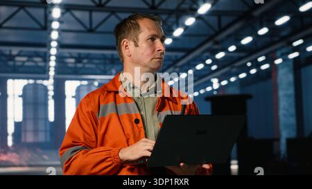 Male worker managing fabrication tasks in a warehouse with laptop to support heavy metal manufacturing process, surrounded by machinery and metal tools for production. Stock Photo