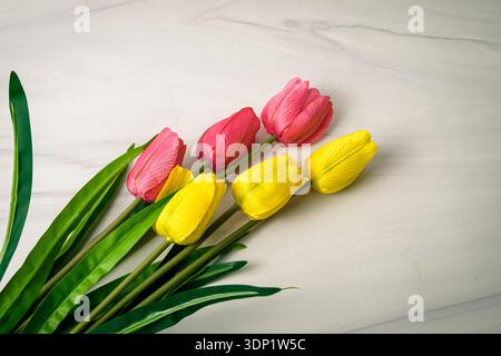 Pink and yellow tulips with green leaves on white marble background, minimal spring flat lay with copy space. Stock Photo
