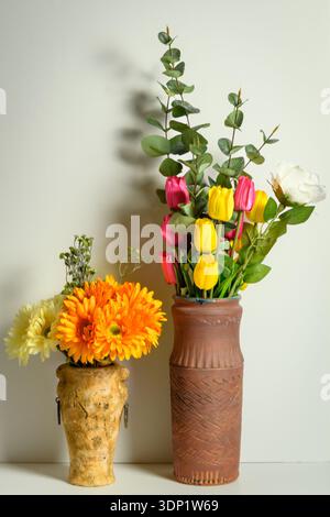 Two rustic vases with tulips, eucalyptus and gerbera flowers against a pale green wall, warm home still life with shadow and copy space. Stock Photo