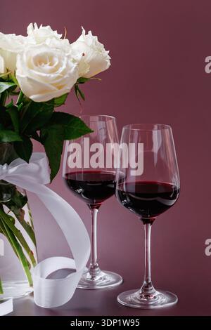 Two Glasses of Red Wine and White Roses on Burgundy Background Stock Photo