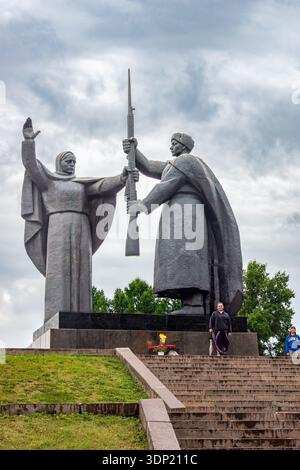 Tomsk, Russia - July 06, 2025. Full-figure composition captures Motherland blessing hand and soldier receiving pose. Eternal Flame burns fnd two visitors at base provide human scale. selective focus Stock Photo