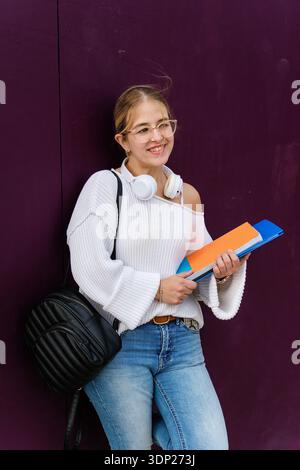 Young woman student smiling and looking away, leaning against a purple wall while holding books and carrying a backpack, representing education and a Stock Photo