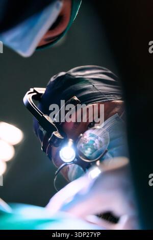 Surgeon In Headlamp Performing Operation Under Bright Surgical Lights In Modern OR During Critical Procedure With Team Stock Photo