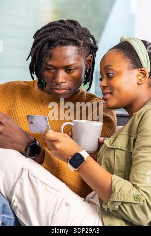 African American couple in brown sweater, green jacket on couch checking smartphone with card, mug Stock Photo