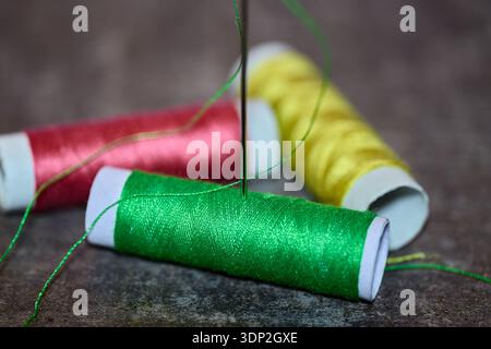 Macro shot of green, red, and yellow thread spools with a needle on a textured surface Stock Photo