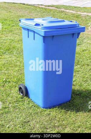 Covered blue plastic garbage container with wheels on the green grass in summer Stock Photo