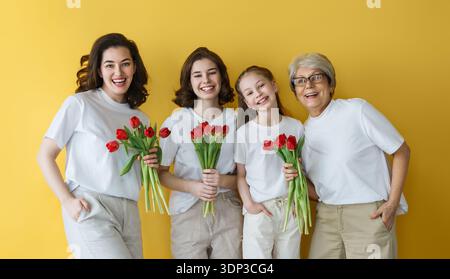 A joyful multigenerational family portrait featuring women and a girl holding vibrant red tulips, symbolizing love and unity. The bright yellow backgr Stock Photo