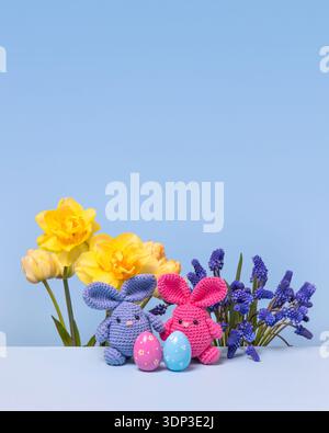 Pink and blue Easter knitted bunnies with multicolored eggs and spring muscari flowers, tulips and daffodils on the blue podium. Concept of Easter Stock Photo