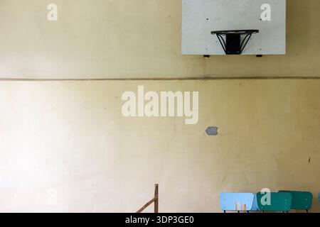Dilapidated indoor basketball court featuring a worn basketball backboard and hoop mounted on a beige wall. The wall displays visible wear and tear, w Stock Photo