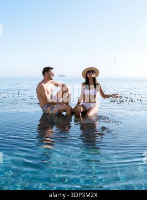 A couple sits in a tranquil pool, soaking in the sun and enjoying the breathtaking ocean scenery of Cape Town. This perfect holiday setting invites relaxation and romance. Stock Photo