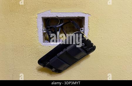Professional electrician repairing a broken black light switch mounted on a yellow textured wall with exposed wires needing urgent technical assistanc Stock Photo