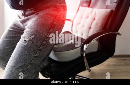 Detail view of person sitting on orthopedic seat cushion placed on office chair, experiencing lower back pain. Red light highlights discomfort area, s Stock Photo