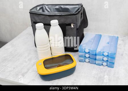Insulated grey cooler bag, two white plastic bottles, blue reusable ice packs, yellow lunchbox on light surface. Objects for keeping food and drinks c Stock Photo