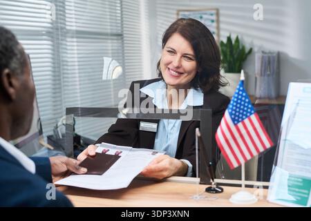 Caucasian middle aged woman smiling while assisting Black man at visa center counter, handing documents and passport across glass partition with American flag visible on desk Stock Photo