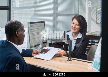 Middle aged Caucasian woman smiling while assisting middle aged Black man submitting documents at visa center reception desk, both engaging in formal application process through glass partition Stock Photo