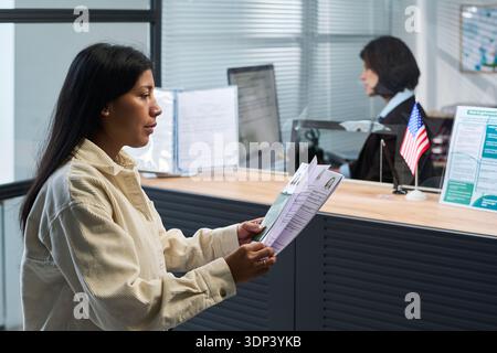 Young adult Hispanic woman holding documents standing at visa center reception counter interacting with middle aged Caucasian woman working behind glass partition, with American flag visible Stock Photo