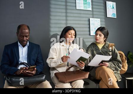 Middle aged Black man using smartphone while two young adult Hispanic women reviewing documents together in visa center waiting area Stock Photo