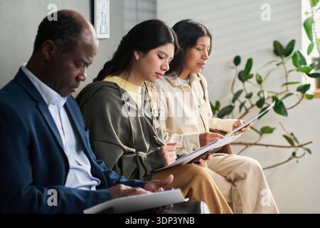 Middle aged Black man and two Hispanic girls sitting in visa center waiting area filling out paperwork, focused on completing application forms Stock Photo
