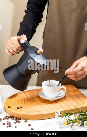Close up of barista hand pouring coffee from black moka pot into white ceramic cup with saucer Stock Photo