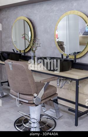 Luxury men's seat in interior of  barber shop.  Comfortable beige leather chair and black hair sink on wooden table with round golden color mirror Stock Photo