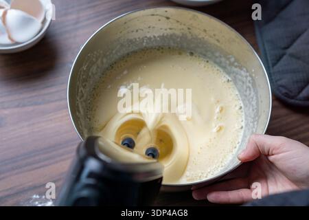 Close-up of cake batter being whipped with an electric hand mixer while a person holds the metal bowl steady. Stock Photo