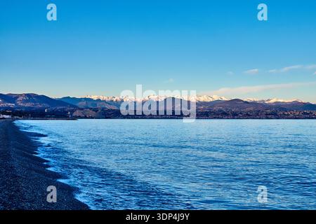 Snow-Capped Mountains Over Calm Coastal Sea With Pebble Beach And Distant Town Under Clear Sky Stock Photo