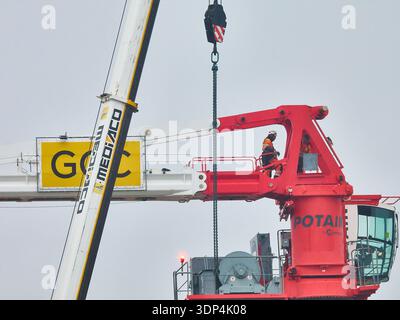 Construction Crane With Workers On Red Tower Crane Platform Lifting Heavy Load In Overcast Weather Stock Photo