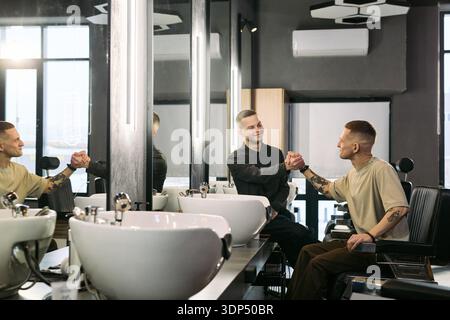 Two young adult Caucasian men greeting each other with handshake in modern barbershop, one sitting in barber chair while other standing and smiling, large mirrors and sinks visible Stock Photo