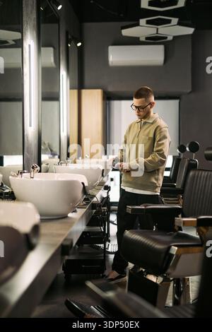 Young adult Caucasian man standing in barbershop preparing electric hair clipper near salon chair and washbasin, focusing on equipment before serving client, modern grooming setting Stock Photo