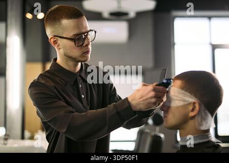 Caucasian young adult man wearing glasses cutting hair of Caucasian young adult man with electric clipper in barbershop, client sitting with eyes covered, barber focusing on haircut Stock Photo
