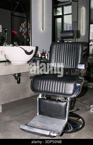 Empty barbershop chair standing in front of mirror and sink with various grooming products on counter, modern professional salon interior prepared for next client Stock Photo