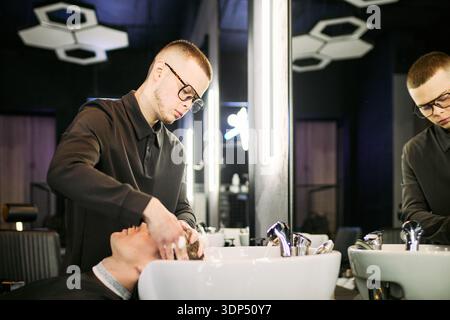 Young adult Caucasian man wearing glasses washing hair of another young adult Caucasian man in barbershop, standing at sink and focusing on hair care, mirror reflecting scene Stock Photo