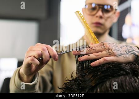 Caucasian young adult man with tattooed hand cutting hair of client using scissors and comb in barbershop, focusing on precise technique and professional grooming service Stock Photo