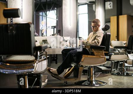 Young adult Caucasian man sitting in barbershop chair using smartphone, waiting for haircut modern grooming salon interior visible in background large mirrors and barber tools on counter Stock Photo