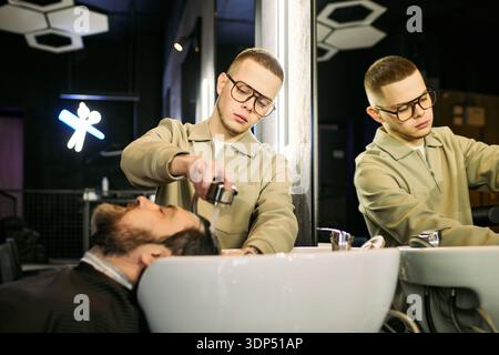 Caucasian young adult man washing hair of bearded Caucasian middle aged man in barbershop, focusing on professional hair care service with both subjects visible in mirror reflection Stock Photo