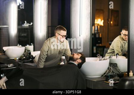 Caucasian young adult man standing over Caucasian young adult man reclining at wash basin in barbershop, barber preparing to wash client's hair, both focused on task, modern salon setting Stock Photo