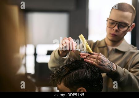 Caucasian young adult man with tattoos cutting hair of young adult man using scissors and comb in barbershop, focusing on precise haircutting technique, hands and tools visible Stock Photo