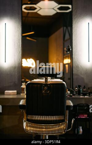 Empty barbershop chair standing in front of large mirror with illuminated wall lights reflecting modern grooming station, various hair styling products arranged on countertop in background Stock Photo