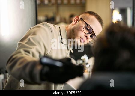 Caucasian young adult man wearing glasses trimming hair of client with electric clipper in barbershop, focused expression showing concentration on precise haircutting technique Stock Photo