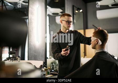 Young Caucasian man wearing glasses cutting hair of seated Caucasian man with beard in modern barbershop, holding electric clipper and comb while focusing on precise grooming Stock Photo