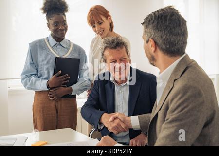 A senior male executive shakes hands with a business partner in a bright office, finalizing a strategic agreement while diverse colleagues look on wit Stock Photo