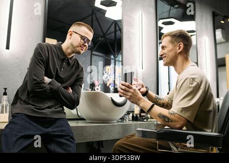 Caucasian young adult man sitting in barbershop chair showing smartphone to Caucasian teenager with short hair and tattoos standing nearby both smiling and interacting Stock Photo