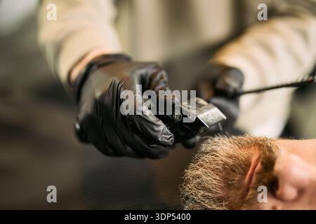Caucasian middle aged man receiving haircut from barber wearing black gloves using electric hair clipper in barbershop, close up on hands and hair, focusing on grooming process Stock Photo