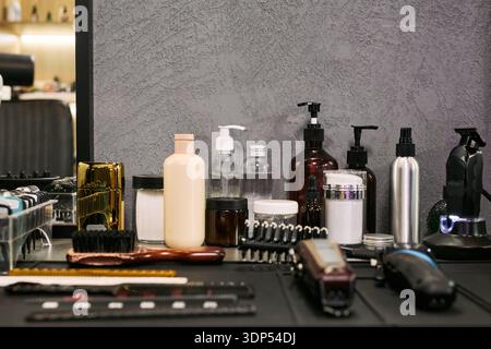 Barbershop countertop displaying various hair care products, grooming tools, electric clippers, combs, brushes, spray bottles, and containers arranged in front of mirror Stock Photo