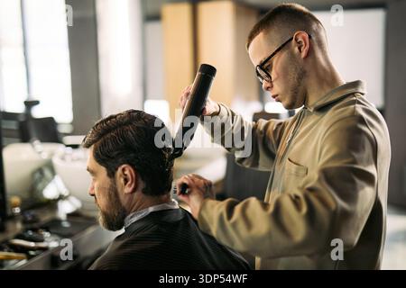 Caucasian young adult man styling hair of Caucasian middle aged man using electric hair clipper in barbershop, focusing on precise haircutting technique and professional grooming service Stock Photo